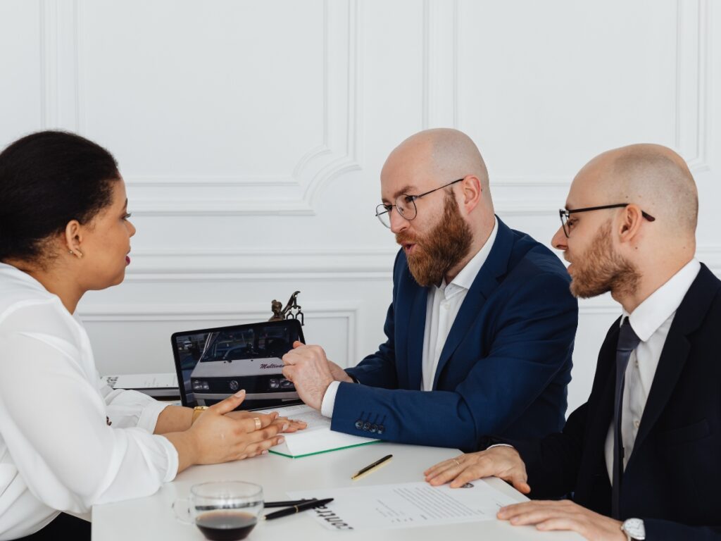 Business Fraud Investigations: How Private Investigators Help Protect Your Company 8 Three people in business attire sit at a table discussing documents, with one person pointing at a laptop screen displaying evidence related to business fraud investigations.