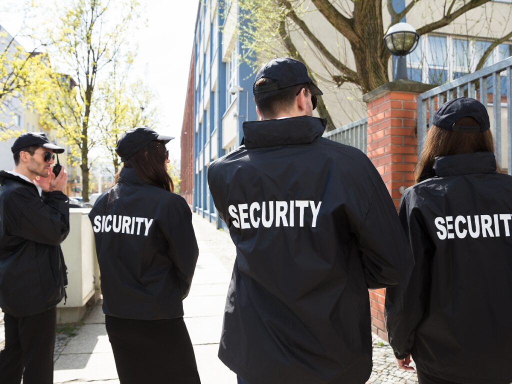 Holiday Event Security: How Security Guards Keep Large Gatherings Safe 1 Four people wearing black jackets and caps labeled "SECURITY" stand outside near a building and a brick wall, providing Holiday Event Security.