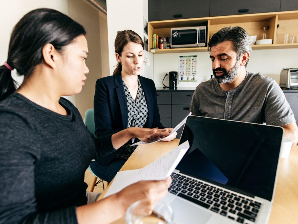 Private Investigator for Legal Cases: Why Lawyers Rely on Professional Investigations 1 Three people sit at a table in an office, reviewing documents with a Private Investigator for Legal Cases. A laptop, papers, and a glass of water are on the table, while shelves and a kitchen area can be seen in the background.