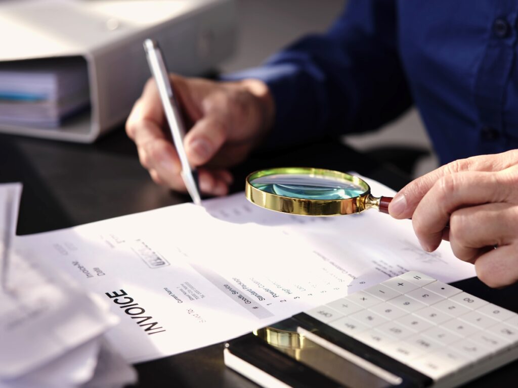 Private Investigator for Legal Cases: Why Lawyers Rely on Professional Investigations 3 A Private Investigator for Legal Cases examines an invoice with a magnifying glass while writing with a pen at a desk filled with documents, a binder, and a calculator.