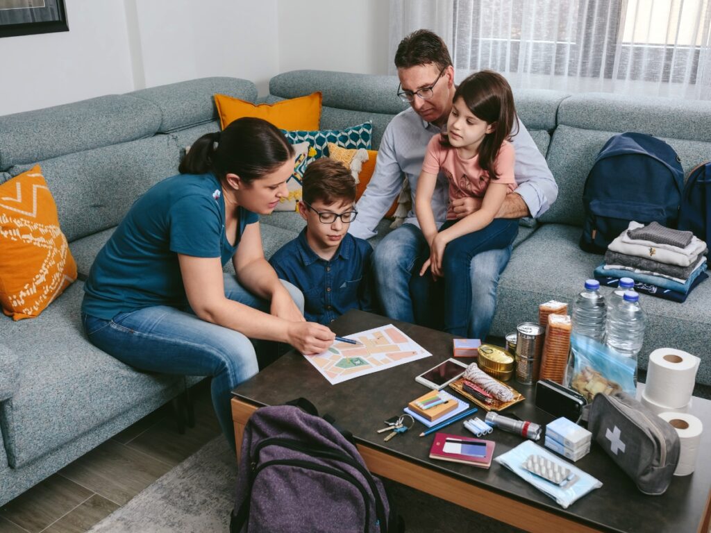 Family Emergency Plan: Prepare for Any Crisis 1 A family sits on a couch, creating their family emergency plan with a map at a table covered with emergency supplies like water, food, a radio, flashlight, and first aid kit.