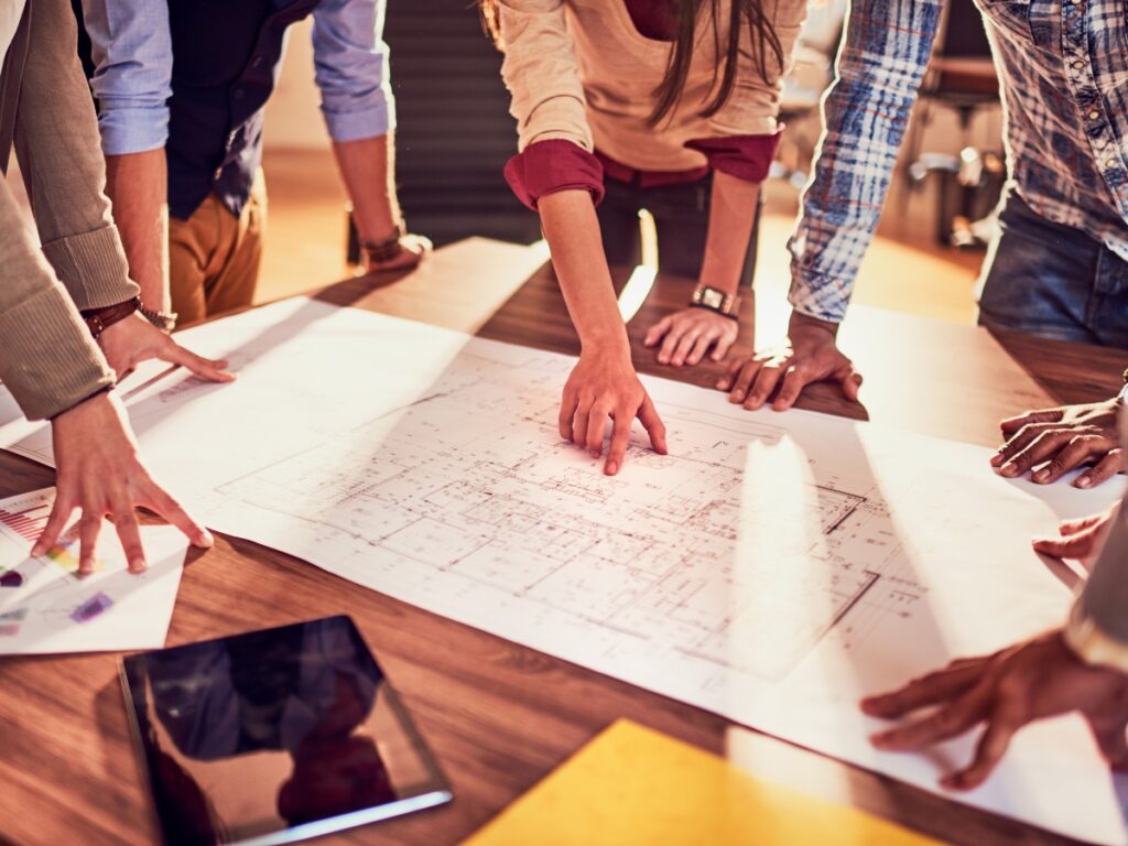 Family Emergency Plan: Prepare for Any Crisis 2 A group of people stand around a table, reviewing and pointing at architectural blueprints or a family emergency plan spread out on the surface.