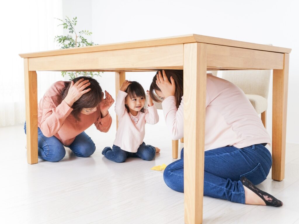 Family Emergency Plan: Prepare for Any Crisis 4 Three people, including a child, are crouched under a wooden table holding their heads, appearing to practice an earthquake safety drill indoors as part of their family emergency plan.