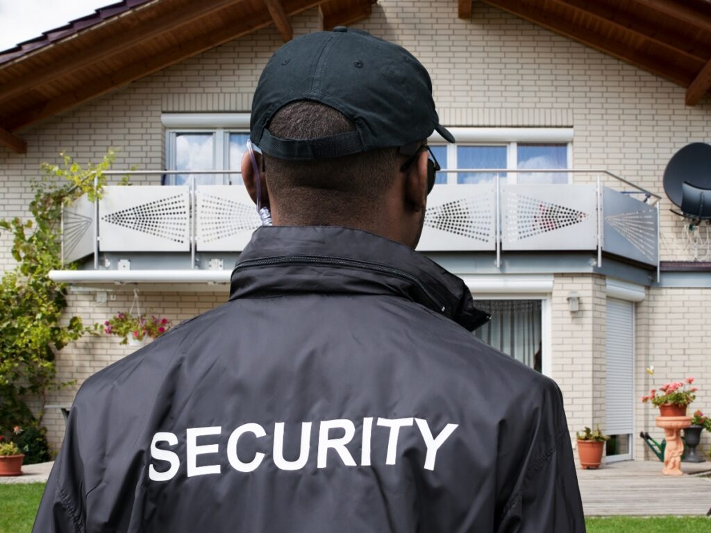 A security guard in a black jacket and cap stands with his back to the camera in front of a residential building, emphasizing the importance of following a home security checklist.
