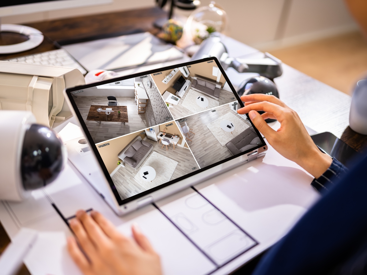 Person monitoring four security camera feeds on a tablet screen at a desk, reviewing a home security checklist, with surveillance equipment and office items visible.